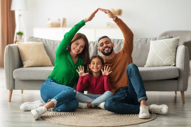 Neşeli Arap ebeveynler, elleri yukarıda yerde mutlu bir şekilde oturan kızlarının üzerinde bir çatı şekli oluştururlar. Aile, yeni evlerinde birlikte vakit geçirmenin keyfini çıkarıyor..