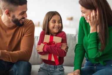 Sıcak bir oturma odasında, Orta Doğulu bir kız gülümseyen ailesinden bir hediye alırken neşeyle ışıldıyor. Birlikte kutluyorlar, aile mutluluğunu ve sevgiyi yakalıyorlar..