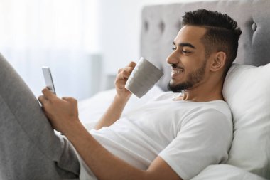 Neşeli genç bir Arap, elinde bir fincan kahveyle yatakta rahatlar ve akıllı telefonunu kullanır. İnternette gezinirken gülümsüyor, gündüzleri evde boş vakit geçiriyor..