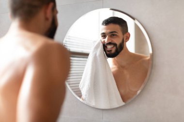 Genç Arap adam modern bir banyoda üstsüz duruyor, beyaz bir havluyla yüzünü siliyor ve aynadaki yansımasına hayran, kendinden emin ve mutlu bir şekilde cilt bakımı yapıyor..