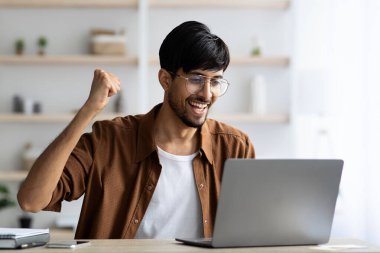 Genç bir adam iyi ışıklandırılmış bir ofiste oturuyor, kutlama için gülümsüyor ve yumruğunu kaldırıyor. Dizüstü bilgisayarıyla mutlu bir şekilde meşgul gibi görünüyor, yaptığı işten zevk alıyor..