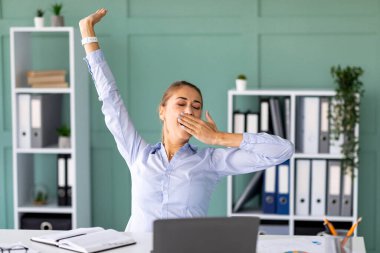 Genç bir bayan modern bir ofiste masasında oturuyor. O bir dizüstü bilgisayar üzerinde çalışırken esniyor ve kollarını esnetiyor. Çalışma alanı raflar ve bitkilerle tasarlanmıştır..