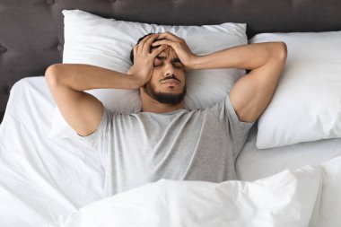 Genç bir Arap yatakta, rahatsızlık ve stres belirtileri gösteriyor. Başını iki eliyle tutuyor, baş ağrısı çekiyor, muhtemelen uykusuzluk ya da akşamdan kalma..