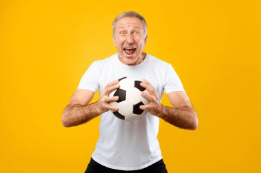 Gri saçlı bir adam futbol topunu iki elinde tutuyor. Kocaman bir gülümsemesi var ve çok mutlu görünüyor. Arka plan parlak sarı, canlı bir atmosfer yaratıyor..