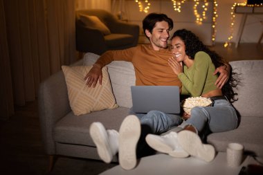 Bir çift kanepede rahatça oturup dizüstü bilgisayarda komedi filmi izlerken birlikte gülüyorlar. Patlamış mısırlarını paylaşıyorlar ve evde rahat bir akşamın tadını çıkarıyorlar. Peri ışıkları rahatlatıcı atmosfere katkı sağlar..