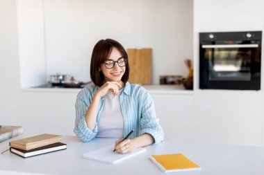 Modern bir mutfakta, gözlüklü genç bir kadın, kitaplarla ve kırtasiyelerle çevriliyken, mutlu bir şekilde bir masaya oturur..