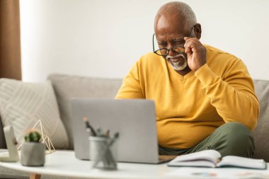 Üst sınıftan bir adam dizüstü bilgisayar çalışırken gözlük takıyor. Kitaplar ve küçük bir bitki ile çevrili, rahat bir oturma odasında bir kanepede oturuyor, evde bir öğleden sonranın keyfini çıkarıyor..