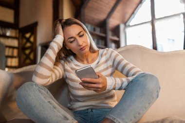Genç bir kadın endişeli bir ifadeyle telefonuna bakarken bacak bacak üstüne atmış bir şekilde kanepede oturuyor. Konforlu odada büyük pencereler ve sıcak ışıklandırma yer alıyor..
