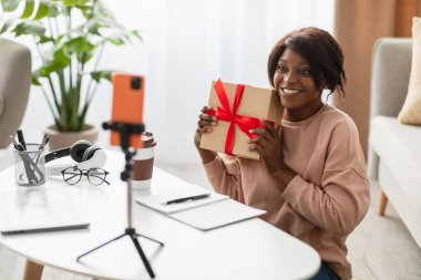 Kırmızı kurdeleli bir hediyeyle masada oturan bir kadın video çağrısına cevap verirken gülümsüyor. Odada bitkiler ve sıcak bir atmosfer var. Paylaşım anları için mükemmel..