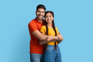 Gülümseyen bir çift, canlı mavi bir arka planda birbirini kucaklıyor. İkisi de gündelik giysiler giyip neşeli ifadeler sergiliyor, mutluluklarını ve bağlantılarını vurguluyorlar..