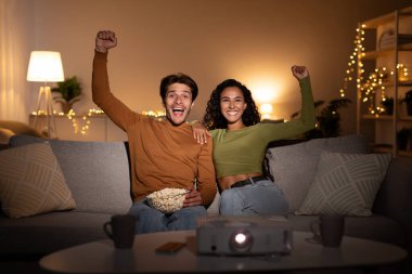 Karı koca, projektör kullanarak spor maçı izlerken sevinçle yumruklarını sallıyor. Birlikte zaferi kutluyorlar, rahat bir oturma odasında kanepede kaliteli zaman geçiriyorlar..