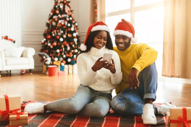 İki kişi bir Noel ağacının yanında yerde oturuyor. Noel Baba şapkası takıyorlar ve akıllı telefona bakıyorlar. Hediyeler etraflarını sarmış, neşeli bir hava yaratıyor..