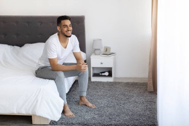 Genç bir Orta Doğulu adam uyandıktan sonra yatağında otururken gülümsüyor..