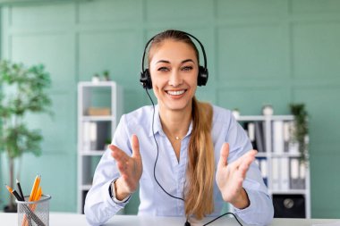 Genç bir bayan ofisinde otururken bir video konferansına katılıyor. Kameraya konuşurken kulaklık takıyor, gülümsüyor ve jestler yapıyor. Çekici bir atmosfer yaratıyor..