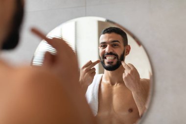 Yakışıklı bir Arap adam banyoda duruyor, aynadaki yansımasını incelerken diş ipi kullanıyor. O, günlük kişisel bakım rutininin bir parçası olarak güçlü dişlerini korur..