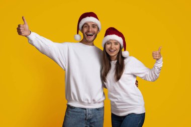 Eşleşen beyaz kıyafetler ve kırmızı Noel Baba şapkaları giyen gülümseyen genç çift, Noel 'i neşeyle kutluyor. Parlak sarı bir arkaplan önünde başparmak işareti ile poz veriyorlar..