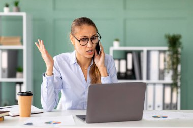 Akıllı telefonuyla konuşan bir kadın, dizüstü bilgisayar programıyla ilgili sıkıntılarını dile getirirken stresli görünüyor. Modern ofis ortamında masasında oturuyor..