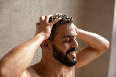 Genç bir Arap erkek model sıcak bir duşun altında durarak mutlu bir şekilde gülümsüyor. Çağdaş bir banyoda başının ve vücudunun üzerinden su taşmasına izin veriyor..