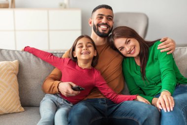 Üç kişilik mutlu bir Arap ailesi televizyon izlerken rahat bir kanepede rahatlar. Gülüşlerini ve gülüşlerini paylaşıyorlar, modern oturma odalarında neşeli anlar yaratıyorlar..