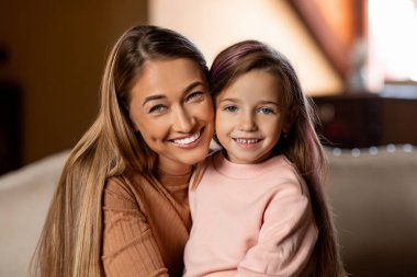 Bir anne ve küçük kızı birbirine yakın, her ikisi de geniş bir gülümseme içinde. İçerideki sıcak ışık, birlikte geçirdikleri zamanların tadını çıkarırlarken sevinçlerini vurguluyor. Sıcak ortam neşe katıyor..