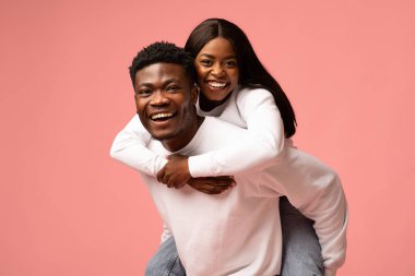 Genç siyah bir kadın erkek arkadaşını sırtında taşıyor, ikisi de kameraya sevinçle gülümsüyor. Neşeli sahne, güçlü bağlarını gösteren yumuşak pembe bir arka planda mutluluklarını yakalıyor..