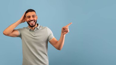 Yüzünde bir gülümsemeyle mavi bir arka plana karşı duran bir adam. Açık gri bir gömlek giyer ve bir tarafı işaret eder, coşku ve neşe gösterir..