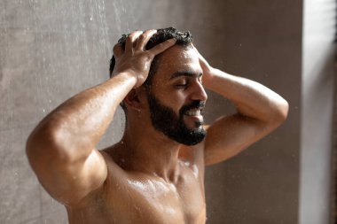Mutlu genç bir Arap erkek evde ferahlatıcı bir duş alıyor. Saçlarını şampuanla yıkarken vücudunun üzerinden su akıyor. Banyoda rahatlatıcı bir atmosfer yaratıyor..