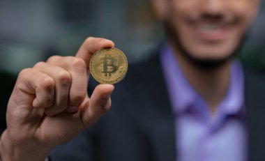 Takım elbiseli bir kişi Bitcoin para tutuyor ve iş ortamında gülümsüyor. Arka planda bulanık ofis elementleri var. Bu da profesyonel bir atmosferi gösteriyor..