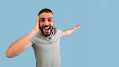 Sakallı genç bir adam mavi bir arkaplana karşı poz verirken geniş bir gülümseme sergiliyor. Heyecan ve neşe ifade ediyor, neşeli bir atmosfer sergiliyor..