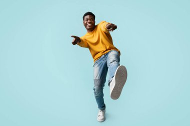 Kaygısız, yakışıklı, komik, bin yıllık siyah adam mavi stüdyo arka planında poz veriyor, kameraya gülüyor ve el kol hareketi yapıyor, Afro-Amerikan genç adam kameraya işaret parmağı ve ayakkabı gösteriyor.