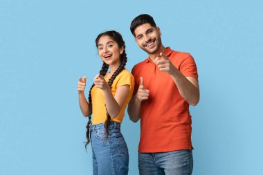 İki genç yetişkin yan yana duruyor, geniş bir gülümsemeyle kamerayı işaret ediyor. Kadın sarı bir bluz giyerken erkek kırmızı bir gömlek giyer. İkisi de neşe saçar..