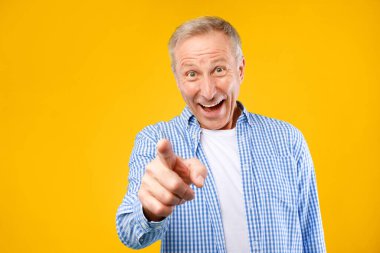 Parlak sarı bir arka planı olan, geniş bir gülümsemeye sahip ve şakacı bir şekilde parmağını gösteren bir adam. Beyaz bir gömleğin üzerine mavi kareli bir gömlek giyer, neşeli bir hava yaratır..