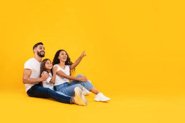 Üç kişilik mutlu bir aile, canlı sarı bir arka planda, yerde oturuyor. Birlikte ilginç bir şeyi işaret ederken gülümsüyorlar ve eğlenceli bir anı paylaşıyorlar..