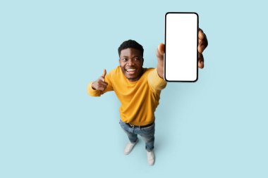 Sarı elbiseli neşeli Afro-Amerikalı genç adam boş ekranlı modern cep telefonunu gösteriyor ve mavi stüdyo arka planında gülümsüyor, en yeni mobil uygulama, üst görünüm, kopyalama alanı öneriyor.