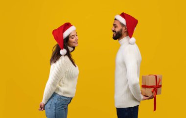 Bir erkek ve bir kadın Noel şapkası giyerken şakalaşıyorlar. Adam arkasında sarılı bir hediye tutuyor, eğlenceli ve şenlikli bir ortam yaratıyor..