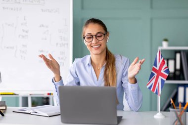 Gülümseyen İngilizce öğretmeni bir online ders veriyor, coşkuyla el kol hareketi yapıyor ve dizüstü bilgisayarındaki webcam 'e bakarken kuralları anlatıyor. Arka planda bir beyaz tahta görünür.
