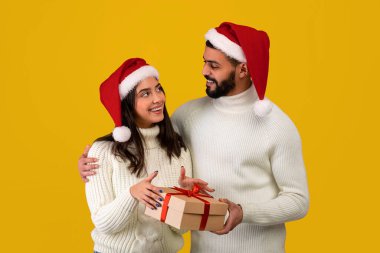 Neşeli bir çift, parlak sarı bir arka planda bir hediye kutusunu tutarken neşe dolu bir anı paylaşıyorlar. Her ikisi de Noel Baba şapkası takıyor ve birbirlerine içtenlikle gülümsüyorlar..
