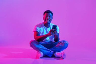 Heyecanlı genç Afrikalı Amerikalı adam neon ışıklarda bağdaş kurarak oturuyor, akıllı telefonuyla mutlu bir şekilde sohbet ediyor ve sosyal medyaya bağlanıyor. Bu canlı ortamda neşe ve coşku yayıyor..