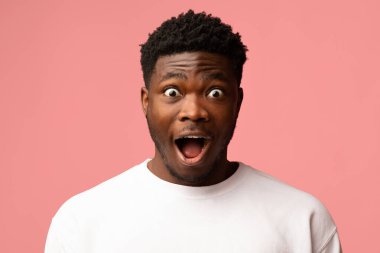 Şaşırmış Afrikalı Amerikalı adam kameraya geniş gözleri ve açık ağzıyla bakıyor. Pembe bir arkaplana karşı tek başına poz veriyor. Parlak bir ortamda güçlü insan duyguları sergiliyor..
