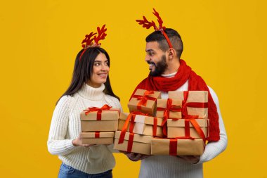 Geyik boynuzu takan mutlu bir çift güzel paketlenmiş hediyelerle birlikte duruyorlar. Mutlu bir anı paylaşıyorlar, bayram sezonunda şenlik ruhunu sergiliyorlar..