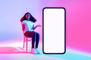 Güzel siyah bayan, boş ekranlı, modern bir akıllı telefonun yanındaki sandalyeye rahatça oturur. Renkli neon ışıklar hareketli bir atmosfer yaratıyor, mobil bir uygulamayı desteklemek için mükemmel..