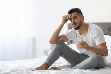 Genç Arap adam kendini kötü hissederek yatakta oturuyor, elinde bir bardak su tutuyor. Kafasına dokunuyor, baş ağrısı belirtileri gösteriyor. Oda aydınlık ve huzurlu. Rahatsızlığını arttırıyor..