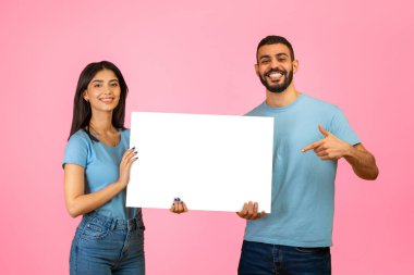 Genç bir çift elinde boş bir tabela tutarken gülümsüyor. Aynı mavi tişörtleri giymişler. Arkaplan canlı bir pembe, yaratıcı fikirler için uygun canlı bir atmosfer yaratıyor..
