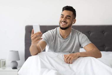 Neşeli genç Orta Doğulu bir adam yatakta oturmuş arkadaşlarıyla video görüşmesi için akıllı telefon kullanıyor. Rahatlamış ve mutlu görünüyor, evdeki zamanının tadını çıkarıyor..