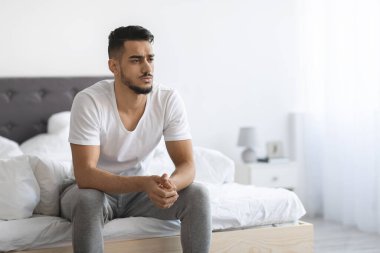 Genç bir Arap, yatağının kenarında dalgın dalgın, endişeli ve derin düşüncelere dalmış bir şekilde oturuyor. Sağlık sorunlarını, özellikle de ereksiyon bozukluğuyla ilgili endişelerini inceliyor gibi görünüyor..