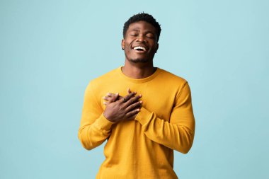 Sarılar içindeki dindar Afrikalı Amerikalı genç adam mavi stüdyo arka planında gözleri kapalı dua ediyor, göğsünü kalbinin üzerine koyuyor ve gülümsüyor, sahneleme alanı olan bir panorama