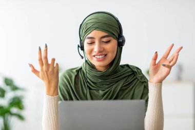 Yeşil tesettürlü genç Arap kadın evdeki dizüstü bilgisayarında bir webinara katılırken kulaklık takıyor. Gülümsüyor ve sohbetten hoşlanıyor gibi görünüyor..