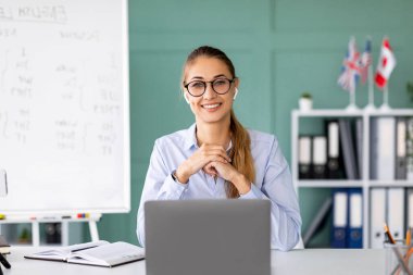 Neşeli bir İngilizce öğretmeni masasında oturuyor, kameraya gülümsüyor ve uzaktan bir ders veriyor. Etrafı dil bilgisi kurallarıyla dolu bir karatahta ile çevrili ve öğrencilerin öğrenmesine yardım etmeye hazır..