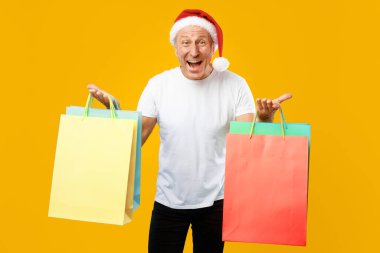 İki elinde de alışveriş torbalarıyla bir adam ayakta duruyor. O bir Noel Baba şapkası takıyor ve gülümsüyor. Arka plan parlak sarı bir renk, neşeli bir atmosfer olduğunu gösteriyor..