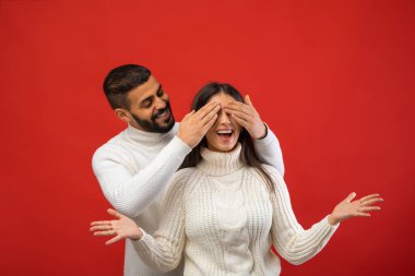 Neşeli bir Arap, elleriyle gözlerini kapatarak kız arkadaşını şaşırtır. O, rahat bir kazakla mutlu bir şekilde duruyor, heyecanını ifade ediyor. Parlak kırmızı arka plan romantik atmosfere katkı yapıyor..
